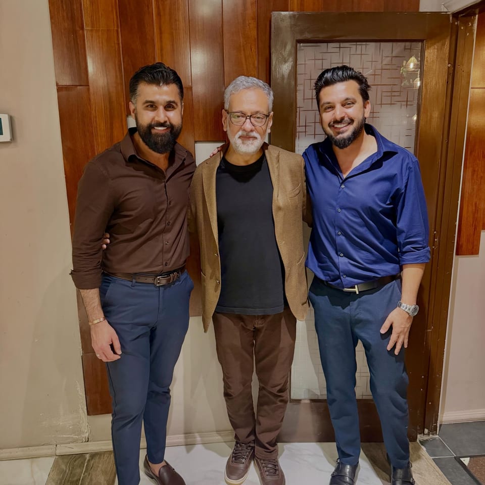 Three smiling men pose together in a warmly lit restaurant hallway with wood-paneled walls and decorative tile.
