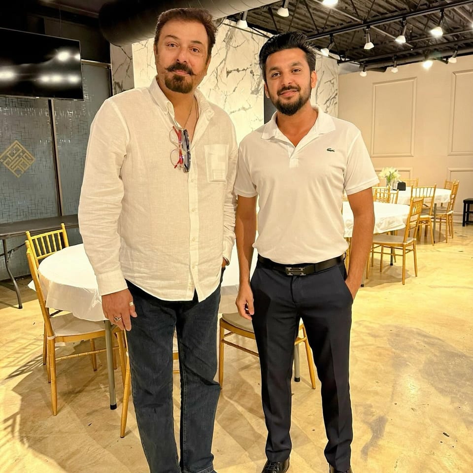Two men pose inside a warmly lit restaurant dining area with white tablecloths, gold chairs, marble walls, and exposed ductwork.