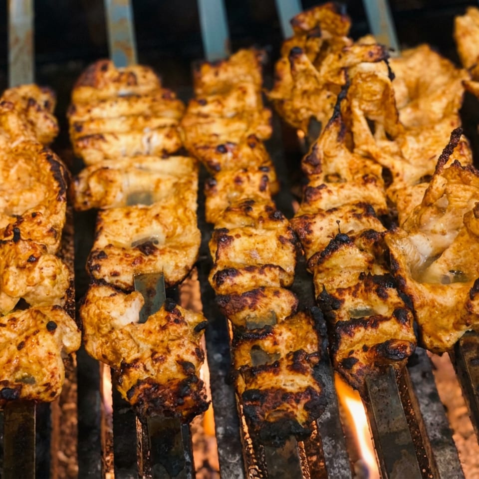 Rows of marinated chicken skewers grill over open flames, showing charred edges and golden-brown seasoning.