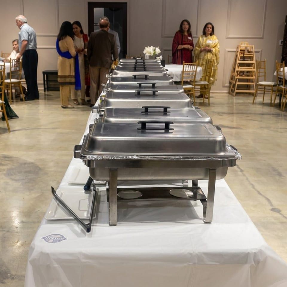 Catered banquet hall with stainless-steel chafing dishes on a long buffet table, surrounded by seated and mingling guests.