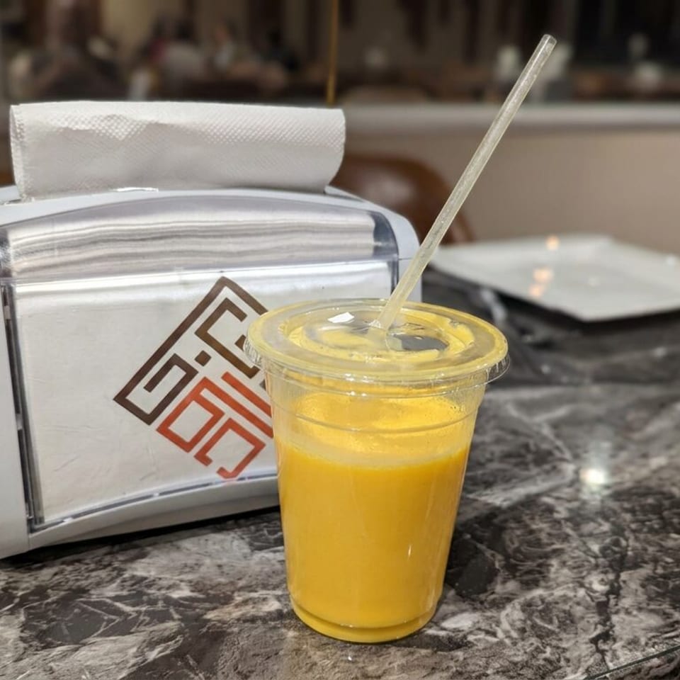 A yellow beverage with a straw sits on a marble restaurant table beside a napkin dispenser.