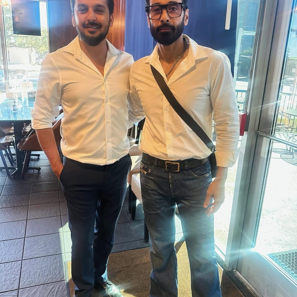 Two men in white shirts pose inside a restaurant near large windows, with tables, chairs, and bright sunlight visible.